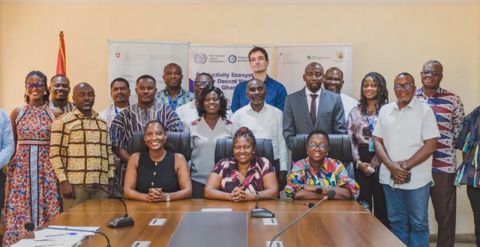 A large group of participants pose around a conference table in front of programme banners and the Ghanaian flag following a partnership meeting in Ghana.