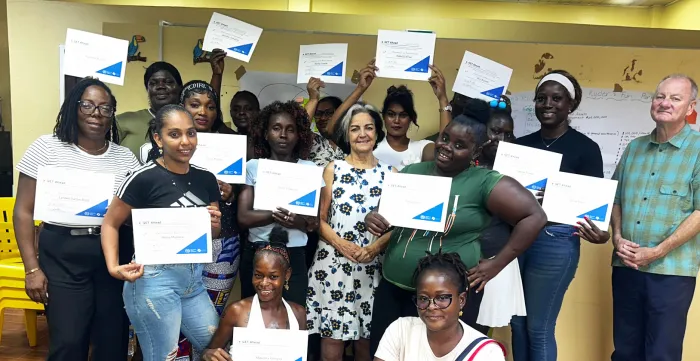A group photograph of approximately 15 individuals, predominantly women, smiling and posing closely together in a brightly lit room with yellow walls. Many of the participants are proudly holding up matching white certificates featuring a blue geometric design in the corner.