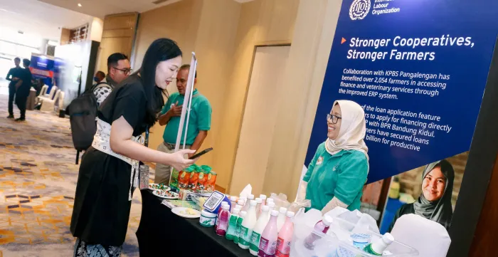 A visitor speaks with a representative of KPBS Pangalengan at a booth displaying dairy products during the Promise II Impact event in Jakarta. An ILO display behind the booth highlights the project’s collaboration with dairy farmers.