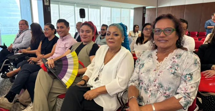 Varias personas sentadas en primera fila miran a cámara; una sostiene un abanico con franjas amarillas, blancas, moradas y negras.