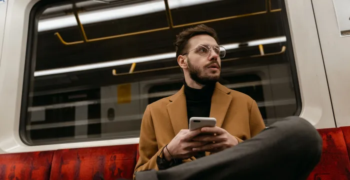 Casually dressed man with eyeglasses holds smartphone on a subway train seat.