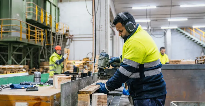 Three men working with wood in a factory, 2 operating machinery and wearing ear defenders and hard hats 