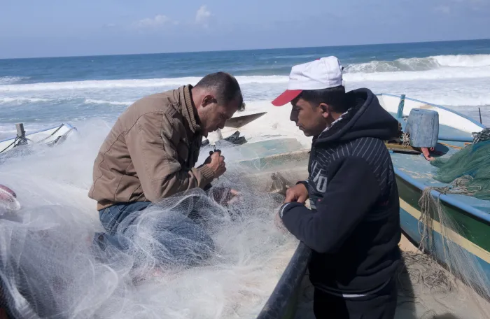 Two fishermen sift through fish net on a boat at the shore, in Gaza