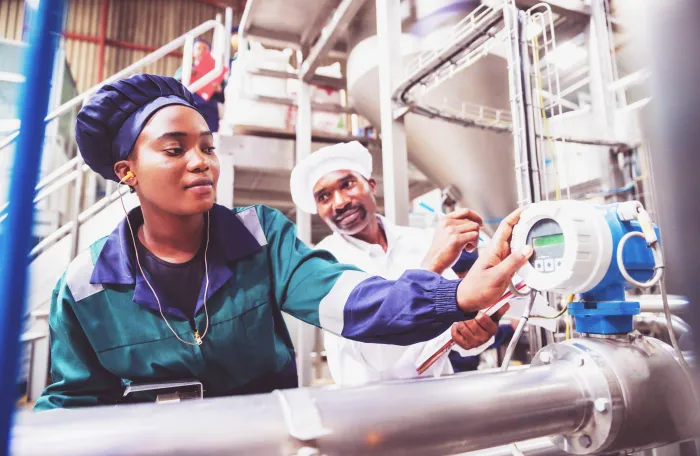 The image depicts two individuals in an industrial setting, where one is adjusting a digital gauge on a large pipe while the other is taking notes.