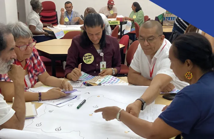Cinco personas adultas sentadas alrededor de una mesa revisan y señalan un mapa sobre papel grande, con marcadores y hojas dispersas.
