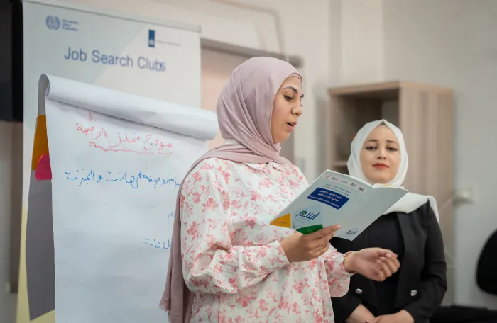 Woman reading from a booklet. In background, a poster says 'Job Search clubs'