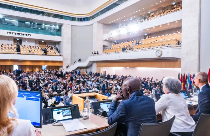 Vista desde la tribuna durante la sesión de apertura de la 114.ª Conferencia Internacional del Trabajo en Ginebra. Las autoridades de la Conferencia están sentadas en la mesa presidencial frente a un auditorio repleto de delegados gubernamentales, de empleadores y de trabajadores, mientras otros participantes ocupan las galerías superiores.