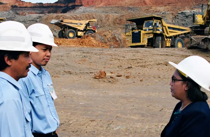 Three people wearing white hard hats stand in conversation at an open-pit mining site.