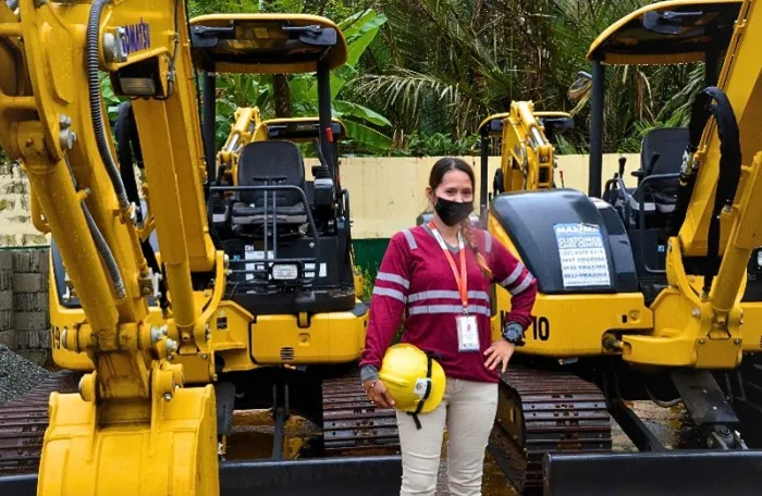 A woman wearing a high-visibility work shirt, face mask, and identification badge stands between two mini excavators while holding a yellow safety helmet.