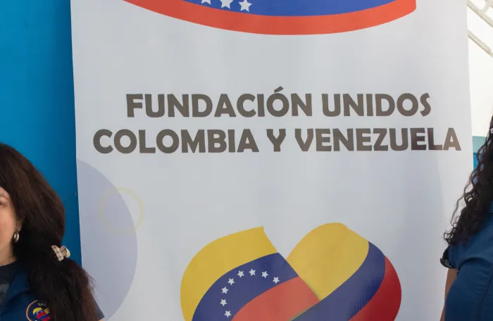 Two female FUNCOLVEN staff members stand smiling beside a banner for Fundación Unidos Colombia y Venezuela, which features the organization's name, logo, and contact information. The photo appears to have been taken indoors during an organizational event or community activity.