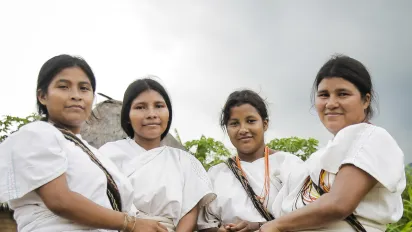 Cuatro mujeres indígenas luciendo su vestimenta tradicional con mantas de color blanco, collares y mochilas terciadas. De fondo una maloca indígena y naturaleza