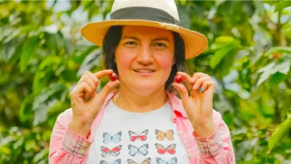 mujer joven con camiseta de mariposas y sombrero aguadeño sostiene con sus dedos dos granos de café, uno en cada mano. En el fondo matas de café