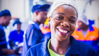 An african woman working in a factory is smiling at the camera, she is wearing a blue and green outfit and hair net, and behind her are more factory workers