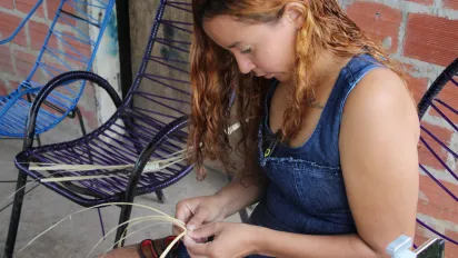 Fátima Laine Sousa Santos está sentada na varanda da frente tecendo a palha  de carnaúba para artesanato. Ela olha para suas mãos, concentrando-se em seu trabalho.