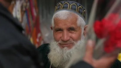 An elderly man with a white beard and a traditional embroidered cap smiles warmly while looking into the camera, framed by blurred figures and flowers in the foreground.