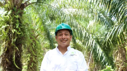 Un hombre posa sonriendo con el fondo de una plantación de palma de aceite. 