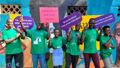 A group of young people in green T-shirts stand in front of a colourful mural, smiling and holding signs calling for innovations to end child labour in their communities.