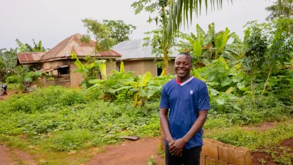 A young Nigerian man, David Chukwuemeka, stands smiling in front of his family’s rural home in Aponmu, Ondo State. He is wearing a blue shirt, surrounded by green plants and simple houses in the background.