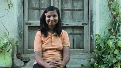 Un portrait de Lata Korde. Elle est assise sur une marche en pierre devant une porte en bois. Des plantes grimpent le long d’un mur bleu de chaque côté des marches. (Inde, 2024)