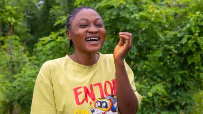 A smiling young woman, Oyefunde Bukola, laughs with her hand raised slightly as she stands outdoors in her community in Ondo State, Nigeria. Lush green foliage surrounds her, and she appears relaxed and joyful.
