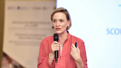 A woman in a pink jacket speaks into a microphone while holding a pen, standing in front of a blurred presentation screen and banner.