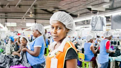 A worker at a garment factory in Haiti looks to camera, representing the ILO's Better Work Haiti programme, which supports improved working conditions in the country's textile and apparel sector. Fellow workers are visible at sewing stations in the background.