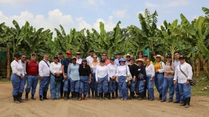 Grupo de 27 personas, compuesto por 13 mujeres y 14 hombres, posan para una foto con las palmas de banano de fondo. La mayoría se encuentra usando gorras o sombreros de ala ancha y todos portan pantalones azules y botas negras de caucho.