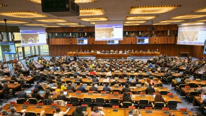 Wide view of the 355th Session of the ILO Governing Body in Geneva. Government, employer and worker representatives are seated in the conference hall facing the dais, while large screens display the proceedings of the meeting.