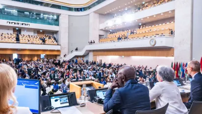 View from the podium during the opening sitting of the 114th International Labour Conference in Geneva. Conference officials are seated at the dais facing a packed assembly hall filled with government, employer and worker delegates, with additional participants seated in the upper galleries. National flags line the conference floor beneath the large auditorium balconies.