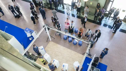 Overhead view of a live recording set at the 114th International Labour Conference in Geneva. Speakers participate in a roundtable discussion on a temporary studio stage while camera operators, technicians and journalists record the conversation. Conference delegates and participants gather around the set, watching, networking and moving through the conference venue.