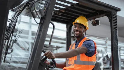 A smiling male worker wearing a yellow hard hat and high-visibility safety vest operating a forklift in an industrial warehouse setting.