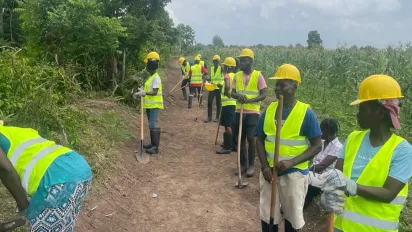 A group of manual laborers wearing yellow hard hats and neon green safety vests work on a dirt path next to a cornfield. Some workers hold shovels and tools while standing or digging along the rural terrain under an overcast sky.