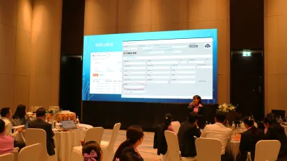 Participants attend a training session in a hotel meeting room, facing a large presentation screen at the front of the hall.