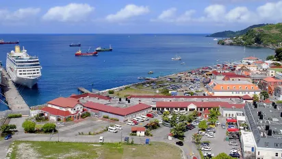 High-angle wide view of St. George’s harbour in Grenada, featuring two large cruise ships docked along the pier in clear blue water. Red-roofed port buildings, parking areas, and the colorful town stretch along the bay toward lush green hills under a bright sky.