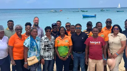 A group of fisheries knowledge-exchange participants standing together on a sandy beach, with bright blue ocean waters and small fishing boats behind them.