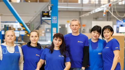 One man and six women workers, including a woman using a walker, stand together on a factory production floor.
