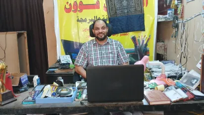 Alhassan Ali sits behind a desk in his mobile phone repair shop. 
