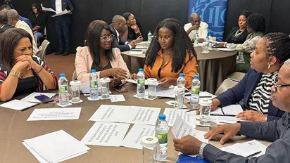 Grupo de pessoas conversam sentados a uma mesa. Na mesa estão papéis, garrafas de água e copos.  