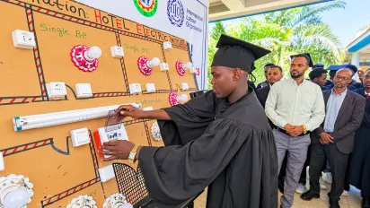 A young man wears a graduation black gown and cap and operates a demonstration electrical panel with other people looking on.