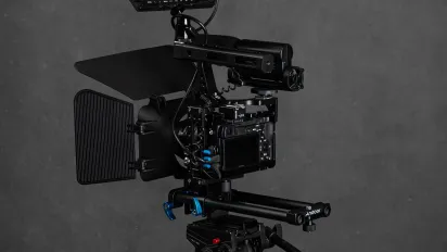 Professional video camera with a monitor on a tripod against a plain grey studio background.