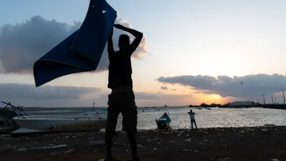 The silhouette of a man lifting a towel over his head, seen against the sunrise, near a lake.