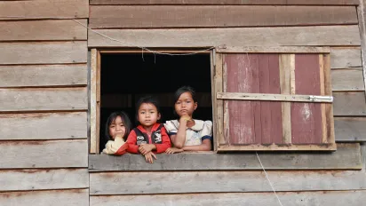 Three children are peeking out of a window in a wooden house.