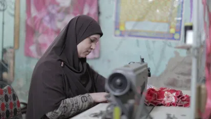 A woman wearing a headscarf sews fabric on a sewing machine in a room with colourful posters and fabric decorations on the walls.