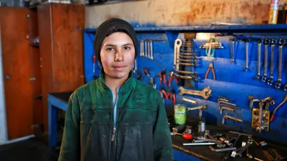 A boy in a green work jacket and black beanie stands confidently in a workshop surrounded by tools neatly arranged on a blue wall.