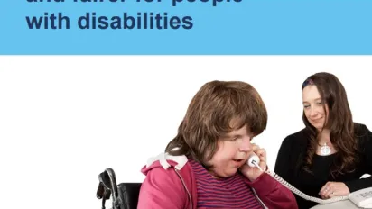 A person using a wheelchair wearing a pink and black striped shirt and pink jacket using a telephone. Another person smiling next to them wearing a black shirt. 