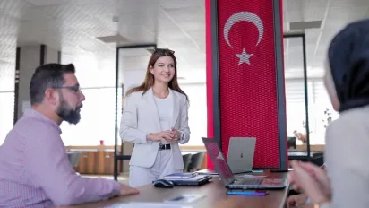 A woman smiles as she listens to two people, against a Turkish flag