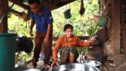 A woman sits behind a big water container. A man stands nearby.