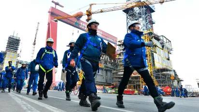 Workers wearing safety gear, including helmets and gloves, walking at a busy industrial site with cranes and construction equipment in the background.