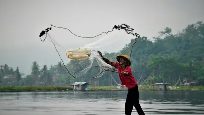 A man in Viet Nam is on a small fishing boat and is throwing a fishing net into the water. He is wearing a red shirt and cone hat.