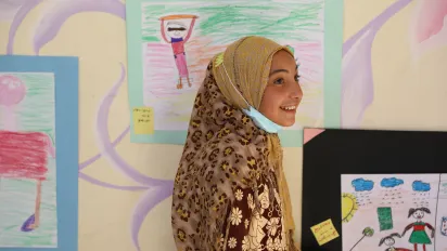 Young girl smiling in front of colourful children's artwork, wearing a patterned headscarf and a mask around her neck.